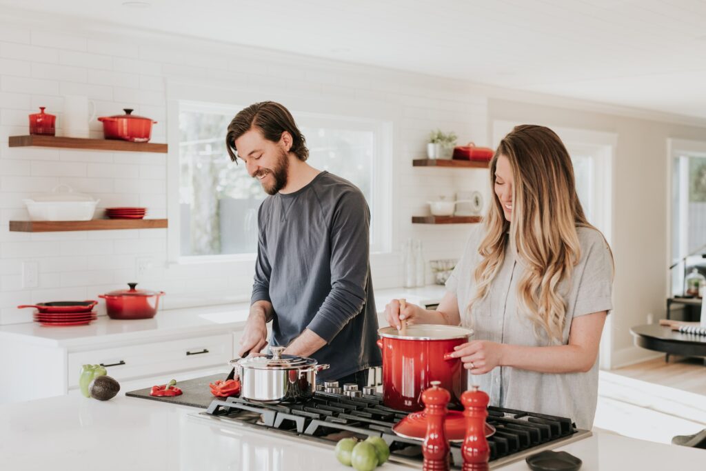 Couple cooking in a newly remodeled kitchen with white cabinets, red kitchenware, and modern appliances.